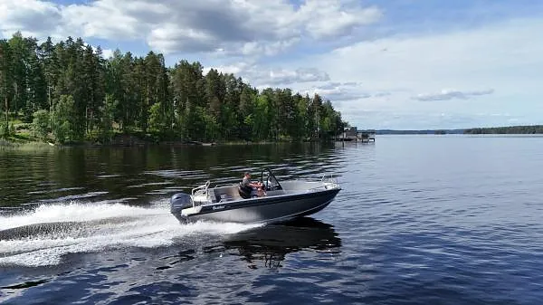 Liten motorbåt i høy fart på stille innsjø, omgitt av skogkledd landskap og spredte skyer.